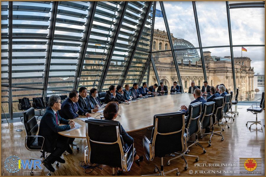 Internationale Delegation an einem großen Konferenztisch unter der Glaskuppel, dahinter das Reichstagsgebäude in Berlin.