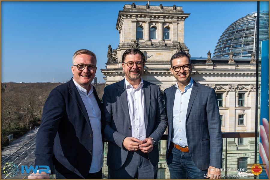 Drei Personen im Gespräch auf einer Terrasse, im Hintergrund das Reichstagsgebäude in Berlin.